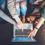 Person typing code prompts into AI chatbot on laptop with mobile phone showing app development