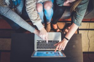 Person typing code prompts into AI chatbot on laptop with mobile phone showing app development